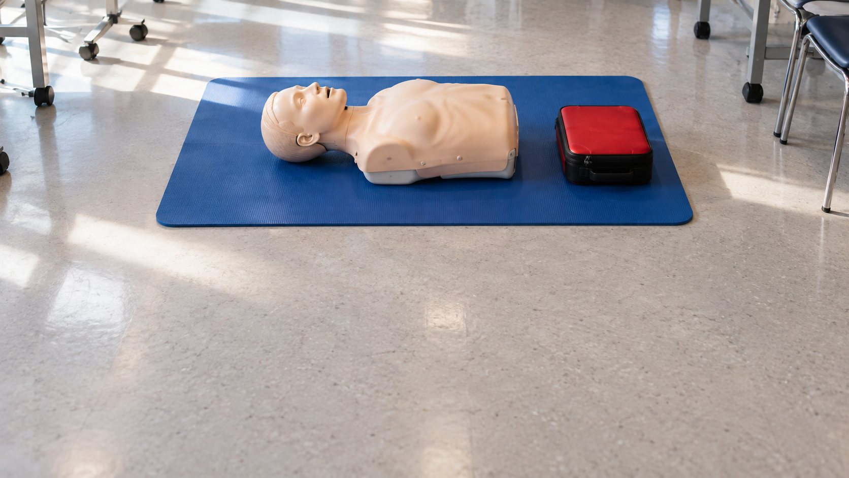 An adult CPR training manikin and a closed training equipment case sit on a mat in a classroom.