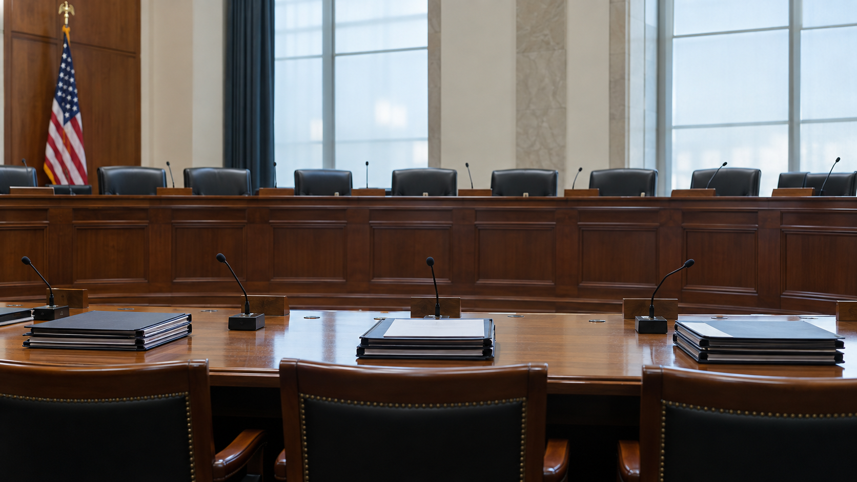 A hearing table with neatly stacked closed folders and microphones before a long empty committee dais.