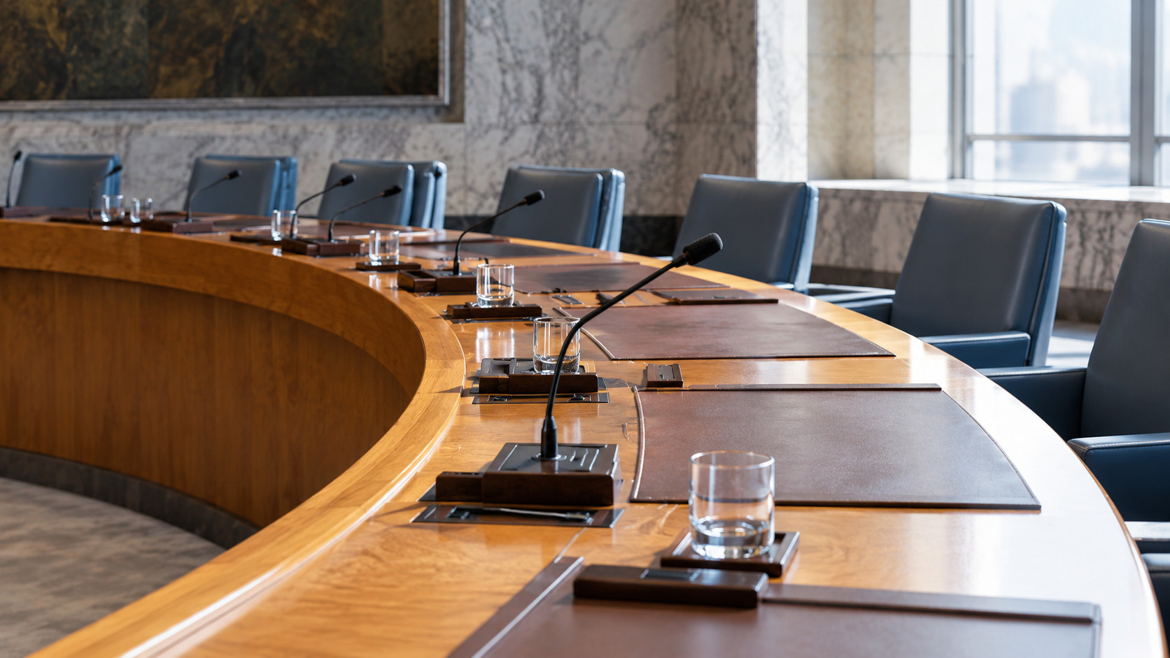 Unoccupied chairs and desk microphones around a curved diplomatic meeting table